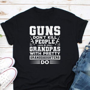 May include: Black t-shirt with white text that reads "GUNS DON'T KILL PEOPLE GRANDPAS WITH PRETTY GRANDDAUGHTERS DO". The shirt features images of guns and bullets. A pair of white sneakers and blue jeans are visible.