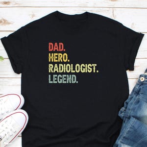 May include: Black t-shirt with the words "DAD. HERO. RADIOLOGIST. LEGEND." in red, yellow, and green vintage-style lettering. White sneakers and blue jeans are visible in the image.