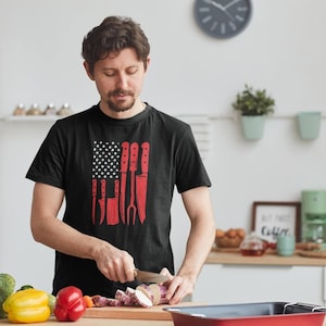 May include: Black t-shirt with a red and white graphic of the American flag, featuring knives and a fork in place of the stripes. The person is cutting vegetables on a wooden cutting board in a kitchen setting.