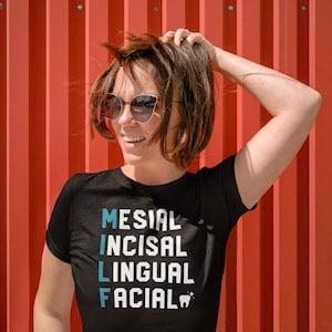 May include: Black t-shirt with the words MESIAL, INCISAL, LINGUAL, and FACIAL in white and blue text. A small tooth graphic is at the bottom. The person in the image is wearing sunglasses and has their hand in their hair.