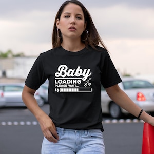 Op de afbeelding: Zwart T-shirt met de witte tekst "Baby Loading Please Wait...". Het ontwerp bevat een laadbalk, hartjes en een babyvoetafdruk. Het T-shirt heeft een ronde hals.