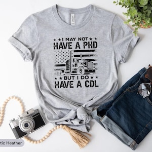 May include: Athletic heather gray t-shirt with the text "I MAY NOT HAVE A PHD BUT I DO HAVE A CDL" and an American flag design with a semi-truck. The shirt is knotted at the waist.