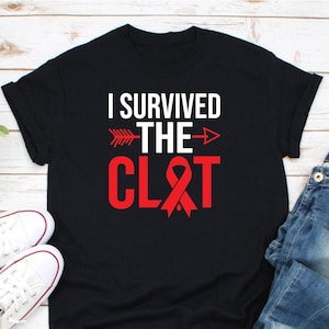 Op de afbeelding: Zwart t-shirt met de tekst "I SURVIVED THE CLOT" in wit en rood. Het ontwerp bevat een pijl en een rood lint. Witte sneakers en een blauwe jeans zijn zichtbaar in de afbeelding.