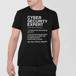 Op de afbeelding: Zwart T-shirt met de witte tekst "CYBER SECURITY EXPERT". De tekst bevat een definitie van een cybersecurity-expert, waaronder "Een persoon die dingen beveiligt die je niet kunt." en "Iemand die precisieschattingen maakt op basis van onbetrouwbare gegevens."
