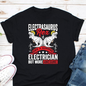 Puede incluir: Camiseta negra con un gráfico que presenta dos ilustraciones de dinosaurios blancos con cascos amarillos. El texto dice "ELECTRASAURUS Rex" encima de los dinosaurios y "LIKE A NORMAL ELECTRICIAN BUT MORE AWESOME" debajo.