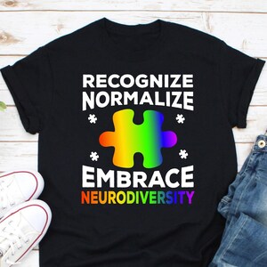 Può includere: T-shirt nero con le parole "RECOGNIZE NORMALIZE EMBRACE NEURODIVERSITY" in bianco e arcobaleno. Un pezzo di puzzle arcobaleno è al centro. Le scarpe da ginnastica bianche sono visibili sul lato sinistro dell'immagine.