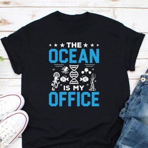 Könnte beinhalten: Schwarzes T-Shirt mit dem Text "THE OCEAN IS MY OFFICE" in Blau und Weiß. Das Design zeigt Illustrationen von Meereslebewesen und eine DNA-Strang. Das Shirt wird mit weißen Turnschuhen und blauen Jeans kombiniert.