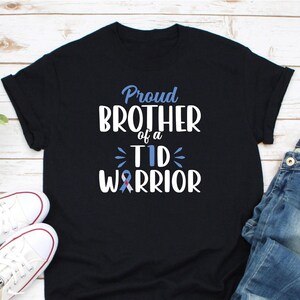 Op de afbeelding: Zwart T-shirt met de tekst "Proud Brother of a TID Warrior" in wit en blauw. Het ontwerp bevat een blauw en rood lint. Witte sneakers en een blauwe spijkerbroek zijn zichtbaar in de afbeelding.