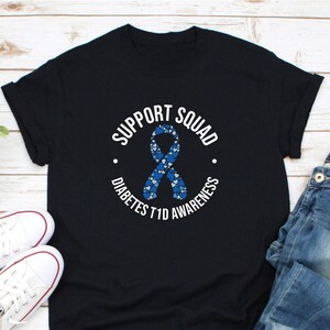 Op de afbeelding: Zwart T-shirt met de tekst "SUPPORT SQUAD" en "DIABETES T1D AWARENESS" in een cirkelvormig ontwerp. Een blauw lint gemaakt van hartvormen staat in het midden. Witte sneakers en een spijkerbroek zijn zichtbaar op de achtergrond.