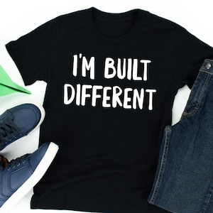 Könnte beinhalten: Schwarzes T-Shirt mit dem weißen Text "I'M BUILT DIFFERENT." Das Shirt wird mit einer blauen Jeans und marineblauen Turnschuhen mit weißen Sohlen kombiniert. Ein grünes Papierflugzeug ist ebenfalls zu sehen.