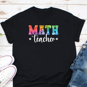 May include: Black t-shirt with the words "MATH Teacher" in a colorful tie-dye design. The word "MATH" is in bold, block letters, and "Teacher" is in a cursive font, with stars on either side. White sneakers and blue jeans are visible.