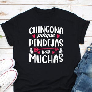 May include: Black t-shirt with white text that reads "CHINGONA porque PENDEJAS hay MUCHAS". The design includes red hearts and an arrow. White trainers and blue jeans are visible in the image.