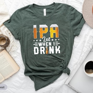 Può includere: Una maglietta verde scuro con la scritta "IPA Lot When I Drink" in un design a tema birra. La maglietta presenta le lettere "IPA" in un font pieno di birra, con una grafica di bottiglia di birra. La maglietta è piegata su una superficie bianca.