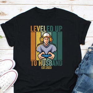 May include: Black t-shirt with a retro design featuring a gamer wearing a headset and holding a controller. The text reads "LEVELED UP TO HUSBAND EST.2023" in orange and white. The design has vertical stripes in orange, yellow, and green.
