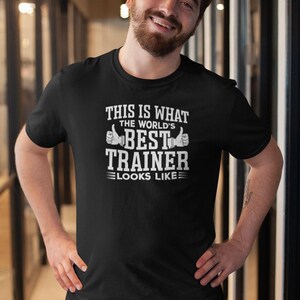 Może przedstawiać: Czarny t-shirt z białym napisem: „THIS IS WHAT THE WORLD'S BEST TRAINER LOOKS LIKE” i grafiką z kciukiem w górę. Koszulka z krótkim rękawem jest noszona przez osobę.