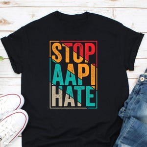 May include: Black t-shirt with the text "STOP AAPI HATE" in a colorful, retro-style design. The text is in a rectangular frame with orange, red, teal, and beige colors. White sneakers and blue jeans are visible in the image.