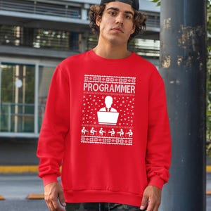Könnte beinhalten: Ein rotes Sweatshirt mit einem weißen Aufdruck, der "PROGRAMMER" und eine Silhouette einer Person am Laptop zeigt, umgeben von Schneeflocken und Figuren an Computern. Das Sweatshirt hat einen Rundhalsausschnitt und ist leger geschnitten.