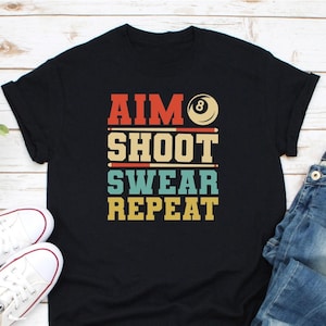 May include: Black t-shirt featuring the text "AIM SHOOT SWEAR REPEAT" in a retro style. The design incorporates a pool ball and cues. White trainers and blue jeans are in the background.