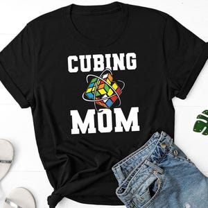 May include: Black t-shirt with the words "CUBING MOM" in white block letters. A colorful Rubik's Cube graphic is centered above the word "MOM". Rolled sleeves and denim shorts are visible.