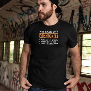 May include: Black t-shirt with the text "IN CASE OF ACCIDENT" in white and orange. The shirt lists instructions: turn off Strava, check bike, and call an ambulance. The person is wearing a black cap and blue jeans.