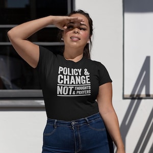 May include: Black t-shirt with white text that reads "POLICY & CHANGE NOT THOUGHTS & PRAYERS." The shirt is paired with blue jeans. The person is outside, with their arm raised to shield their eyes from the sun.