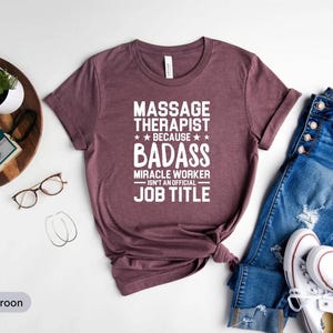 Op de afbeelding: Athletic maroon t-shirt met de witte tekst "MASSAGE THERAPIST BECAUSE BADASS MIRACLE WORKER ISN'T AN OFFICIAL JOB TITLE". Het shirt wordt gecombineerd met een blauwe jeans en witte sneakers.