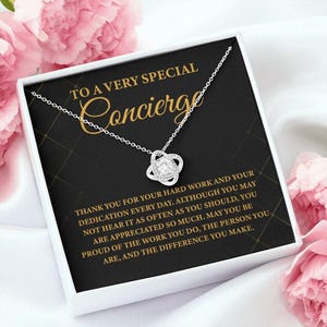 Peut inclure: Un collier argenté avec un pendentif en forme de cœur, présenté dans un coffret blanc. Le coffret porte l'inscription "TO A VERY SPECIAL Concierge" et un message de remerciement. Des fleurs roses encadrent le coffret.