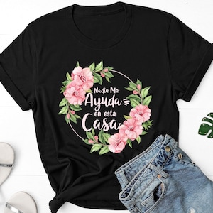 May include: Black t-shirt with a floral wreath design and the text "Nadie Me Ayuda en esta Casa" in white script. The wreath features pink flowers and green leaves. The shirt is paired with denim shorts, red sunglasses, and a green leaf keychain.