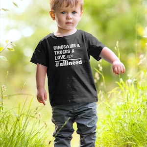 May include: A young child wearing a black t-shirt with the text "DINOSAURS & TRUCKS & LOVE. #allineed" in white. The child is also wearing gray pants and light-colored shoes. The photo is taken outdoors with a blurred green background.