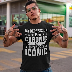 Puede incluir: Camiseta negra con texto blanco: "MY DEPRESSION IS CHRONIC BUT THIS ASS IS ICONIC". La camiseta es de cuello redondo. La persona que lleva la camiseta tiene tatuajes en brazos y manos.