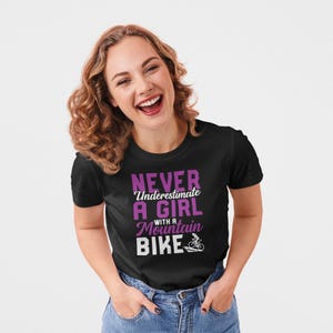 Può includere: T-shirt nero con la scritta "NEVER Underestimate A GIRL with a Mountain Bike" in viola e bianco. La maglietta presenta una grafica di un ciclista su una mountain bike. La modella indossa jeans blu.