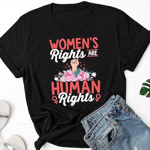 Puede incluir: Camiseta negra con el texto "WOMEN'S RIGHTS ARE HUMAN RIGHTS" en blanco y rojo. El diseño incluye un puño en alto, flores rosas y el símbolo femenino. La camiseta se combina con pantalones cortos de mezclilla, gafas de sol rojas y un llavero verde en forma de hoja.