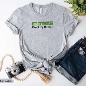 May include: Athletic heather gray t-shirt with the text "sudo rm -rf* Don't try this at~". The shirt is styled with denim shorts, sunglasses, and a vintage camera. A string of wooden beads and a small plant are also in the image.