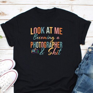 May include: Black t-shirt with the text "LOOK AT ME Becoming a PHOTOGRAPHER & Shit" in colorful letters. A graphic of a camera is included. White sneakers and blue jeans are visible in the image.