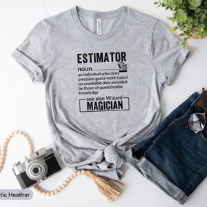 May include: A grey t-shirt featuring the word "ESTIMATOR" and its definition in black text. The definition humorously describes an estimator as someone who does precision guess-work. The shirt is styled with denim shorts and sunglasses.