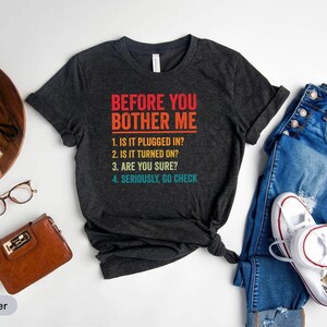 Puede incluir: Camiseta gris oscuro con el texto "BEFORE YOU BOTHER ME" y una lista de preguntas. La camiseta se combina con vaqueros azules y zapatillas blancas. Otros accesorios incluyen una cámara, gafas y una cartera marrón.