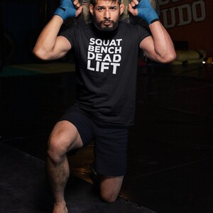 May include: Black t-shirt with the text "SQUAT BENCH DEAD LIFT" in white. The person is wearing black shorts and blue hand wraps, holding a camouflage sandbag. The background is a gym setting.