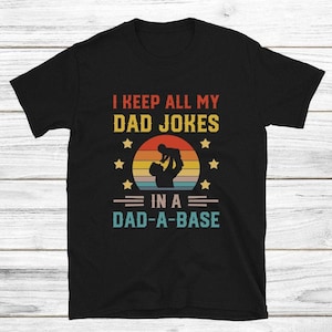 Puede incluir: Camiseta negra con la frase "I KEEP ALL MY DAD JOKES IN A DAD-A-BASE". El diseño muestra la silueta de una persona sosteniendo a un niño, con un fondo de puesta de sol y estrellas.