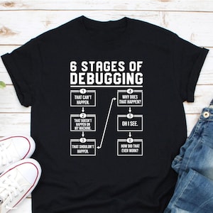 May include: Black t-shirt with a white graphic illustrating the "6 Stages of Debugging." The stages include phrases like "That can't happen" and "Oh I see." The shirt is paired with blue jeans and white sneakers.