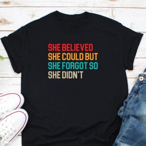 May include: Black t-shirt featuring the quote "SHE BELIEVED SHE COULD BUT SHE FORGOT SO SHE DIDN'T" in red, orange, teal, and beige. The shirt is styled with white trainers and blue jeans.