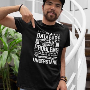 以下が含まれることがあります： 「I'M A DATABASE SPECIALIST I SOLVE PROBLEMS YOU DON'T KNOW YOU HAVE IN WAYS YOU CAN'T UNDERSTAND」と白文字で書かれた黒いTシャツ。人物は黒いキャップと青いジーンズを着用しています。