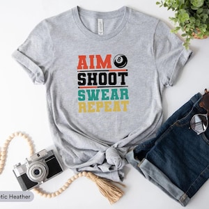 May include: Grey athletic heather t-shirt featuring the text "AIM SHOOT SWEAR REPEAT" in red, black, teal, and yellow, with a pool ball graphic. The shirt is paired with blue denim shorts and sunglasses.