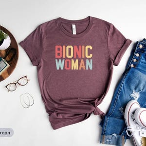 May include: Maroon t-shirt with the words "BIONIC WOMAN" in a retro font. The shirt is styled with blue denim jeans, white trainers, and accessories on a white background.