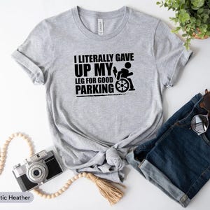 May include: Athletic heather gray t-shirt with the text "I literally gave up my leg for good parking" in black. The graphic includes a person in a wheelchair. The shirt is paired with denim shorts and sunglasses.