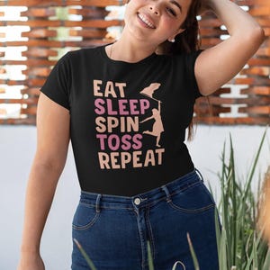 May include: Black t-shirt with the words "EAT SLEEP SPIN TOSS REPEAT" in pink and peach lettering. A silhouette of a person tossing a frisbee is next to the text. The person is wearing blue jeans.