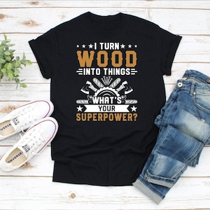 Op de afbeelding: Zwart t-shirt met de tekst "I TURN WOOD INTO THINGS. WHAT'S YOUR SUPERPOWER?" in wit en bruin. Het ontwerp bevat bijlen, een zaagblad en sterren. Witte sneakers en een blauwe jeans zijn ook zichtbaar.
