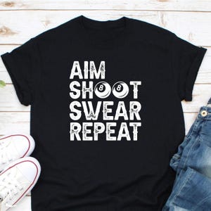 May include: Black t-shirt with the white text "AIM SHOOT SWEAR REPEAT". The word "SHOOT" features two white pool balls with the number 8. White canvas trainers and blue jeans are visible in the image.