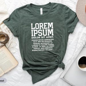 Puede incluir: Una camiseta verde bosque con el texto "LOREM IPSUM DOLOR SIT AMET" en blanco. La camiseta está anudada a la cintura. Un sombrero, una taza de café, un reloj y un libro abierto también están en la imagen.