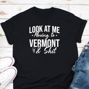 Könnte beinhalten: Schwarzes T-Shirt mit weißem Text, der lautet: "LOOK AT ME - Moving to - VERMONT & Shit." Das Shirt zeigt auch eine kleine Grafik der Umrisse des Bundesstaates Vermont. Weiße Turnschuhe und blaue Jeans sind im Bild zu sehen.