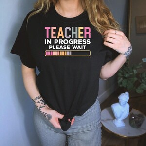 Może przedstawiać: Czarny t-shirt z napisem "TEACHER IN PROGRESS PLEASE WAIT" w kolorze różowym, białym i żółtym. Koszulka posiada grafikę paska ładowania. Koszulka jest zawiązana w pasie.